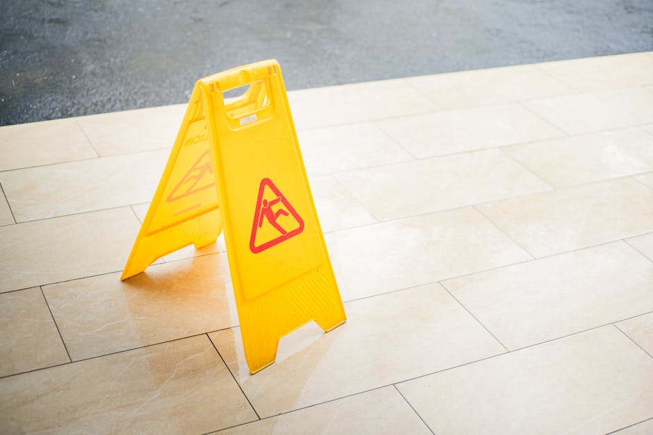 A yellow caution wet floor sign placed on a beige tiled floor near a clean, well-lit doorway or corridor, warning of a potential slip hazard. The sign features a red triangle with a black figure slipp