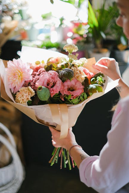A person holding a large bouquet of fresh flowers in their hands, featuring pink carnations, soft pink dahlias, white and purple statice, and green foliage. The bouquet is wrapped in cream-colored pap