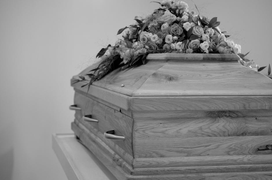 A wooden funeral casket with smooth, polished surface and visible wood grain, positioned on a white stand against a plain light-colored wall. On top of the casket is a large floral tribute composed of