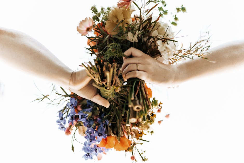 A pair of hands holding a vibrant floral bouquet, featuring an array of fresh flowers including orange poppies, light pink roses, blue delphiniums, white peonies, and various greenery. The bouquet is