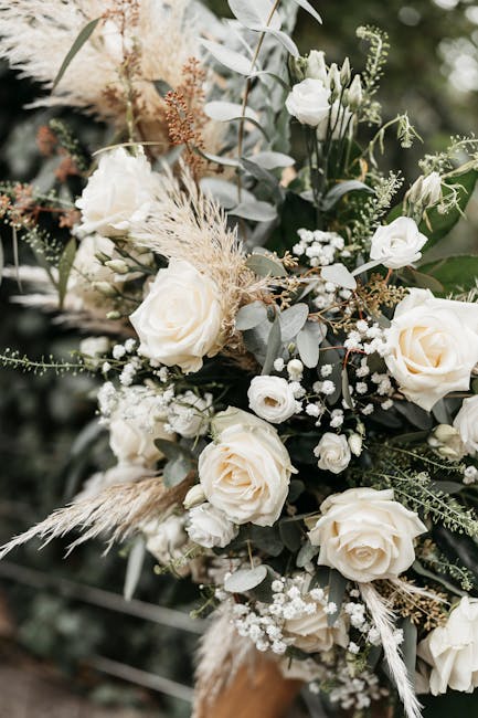 A lush floral arrangement featuring creamy white roses, accentuated by sprigs of baby's breath, silver eucalyptus leaves, and delicate dried grasses, arranged in a natural, cascading style. The flower