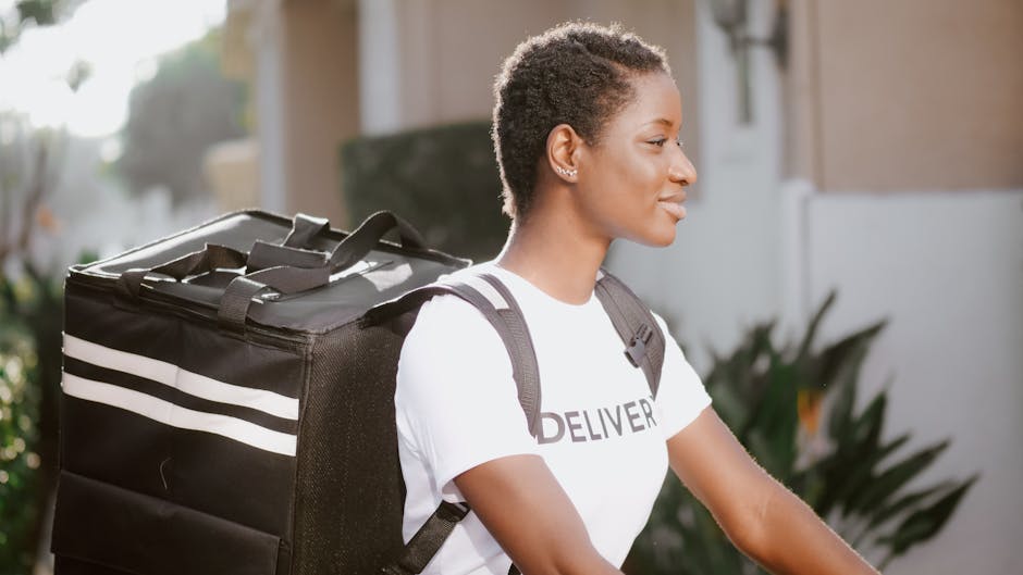 A smiling female delivery courier with short, natural hair, wearing a white t-shirt printed with 'DELIVER' and carrying a large black insulated delivery bag with white stripes, standing outdoors durin